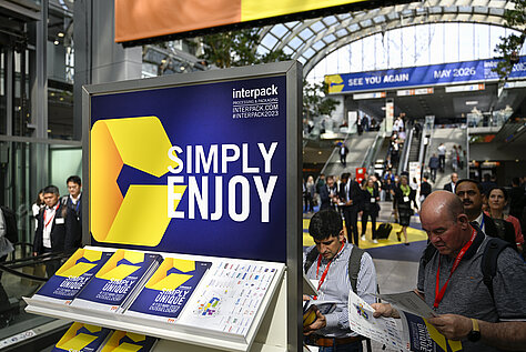 Um estande da Interpack Alliance em uma feira comercial, com um grande letreiro azul e amarelo com os dizeres “SIMPLY ENJOY”. Folhetos com o logotipo estão expostos em um suporte, e visitantes podem ser vistos ao fundo do evento.