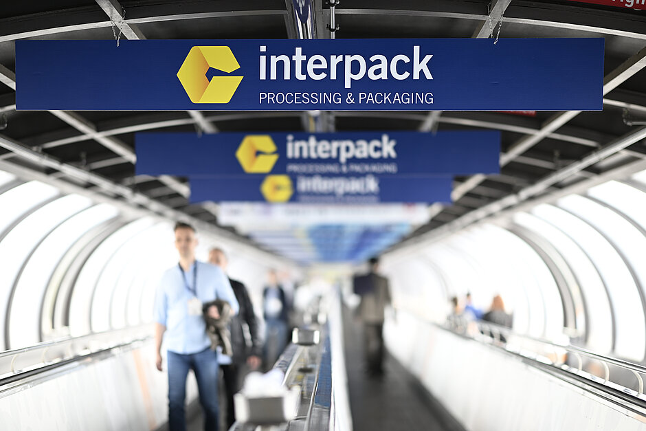 Pessoas caminhando em uma esteira rolante sob várias placas azuis com o logotipo amarelo da interpack e o texto “interpack PROCESSING & PACKAGING” em uma estrutura semelhante a um túnel.
