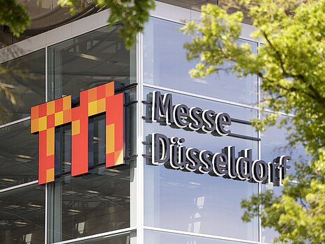 O exterior de um edifício moderno de vidro com o logotipo “Messe Düsseldorf” em destaque na fachada, parcialmente emoldurado por galhos de árvores verdes.