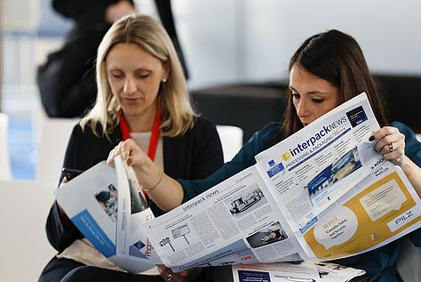 Duas mulheres sentadas, lendo uma edição do jornal “interpack news”, concentradas no conteúdo relacionado ao evento.