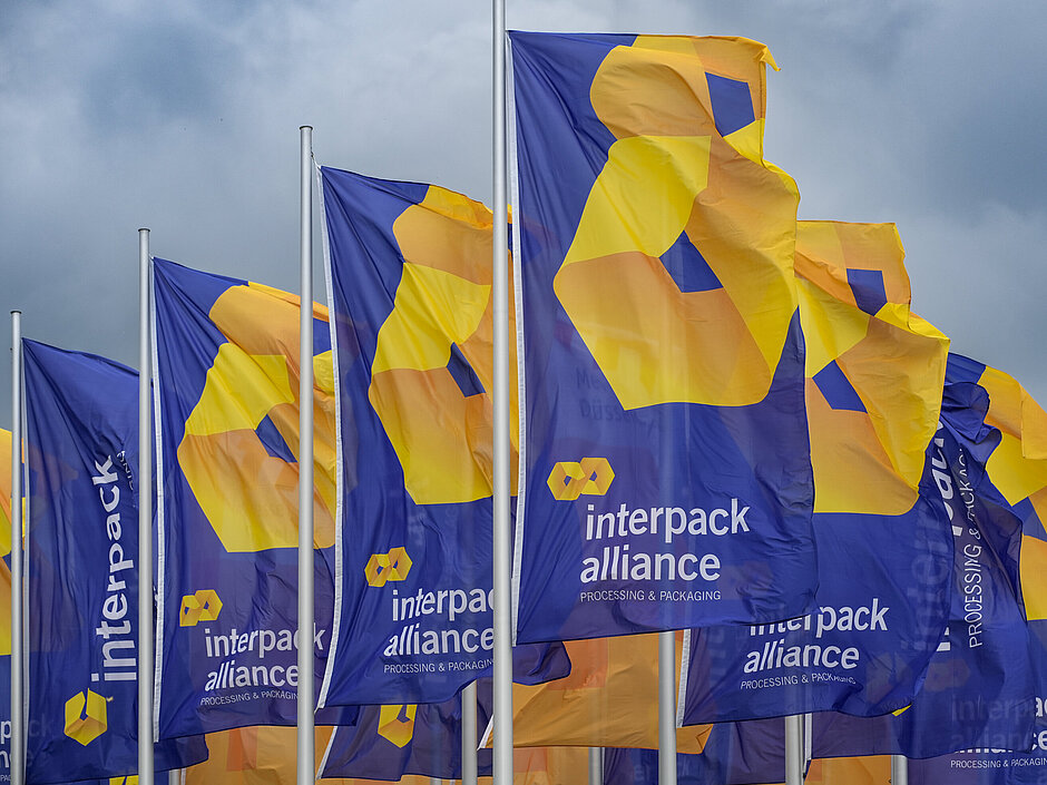 Várias bandeiras azuis tremulando, exibindo o logotipo amarelo-alaranjado e o texto “interpack alliance – Processing & Packaging” contra um céu nublado.
